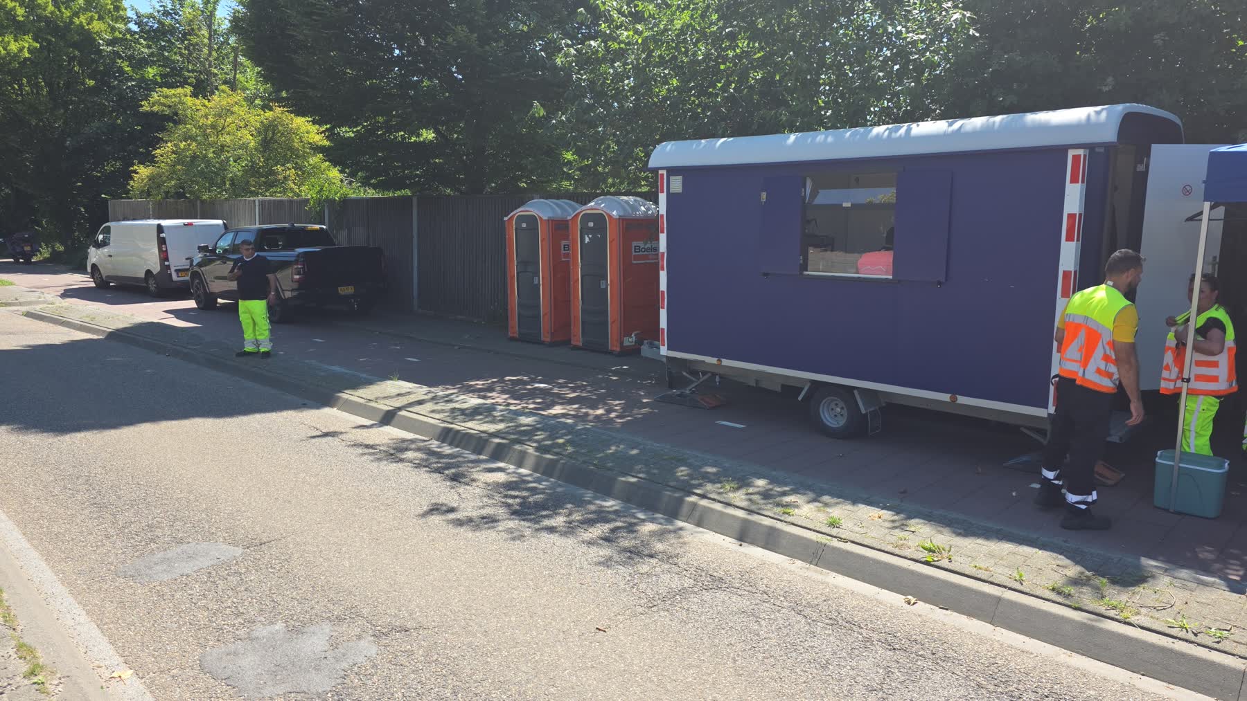 Werklocatie met schaftkeet, sanitaire units en bedrijfswagens langs de weg, met medewerkers in hesje